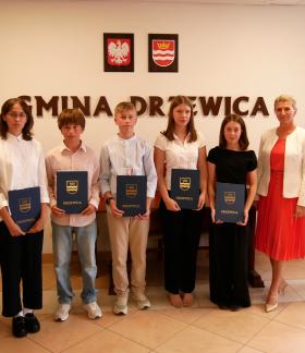 Pozowane zdjęcie grupowe w sali konferencyjnej Urzędu Miejskiego w Drzewicy. Pięcioro uczniów trzymających niebieskie pamiątkowe teczki stoi w centrum. Po bokach Pani Dyrektor ZEAS i Burmistrz Drzewicy. W tle widoczny napis „GMINA DRZEWICA” oraz godło i herb.
