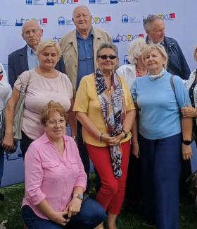 Grupa kilkunastu seniorów reprezentujących Gminę Drzewica pozuje do pamiątkowej fotografii w plenerze na tle promocyjnej ścianki Województwa Łódzkiego podczas Wojewódzkich Obchodów Dnia Seniora