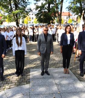  Pięć osób w strojach galowych – samorządowcy i uczennica SP w Drzewicy - stoi w szeregu w parku wyłożonym kostką przed Pomnikiem Pamięci w Drzewicy. W tle widać zgromadzoną młodzież szkolną, poczet sztandarowy i drzewa.