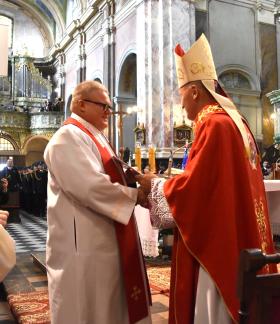Uroczystość kościelna we wnętrzu świątyni w Studziannie. Biskup Marek Solarczyk w czerwonej szacie liturgicznej i mitrze wręcza akt nominacji ks. Grzegorzowi Włodarczykowi w białej albie i czerwonej stule. Z lewej strony przygląda się inny duchowny. W tle zabytkowe organy, nawy kościoła oraz zgromadzeni wierni i strażacy. 