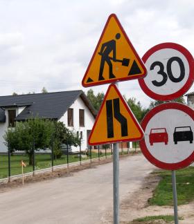 Znaki drogowe ostrzegające o robotach drogowych, zwężeniu jezdni, ograniczeniu prędkości do 30 km/h oraz zakazie wyprzedzania, ustawione przy remontowanej drodze w sąsiedztwie domów jednorodzinnych.