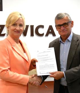 Pani Dyrektor SP w Radzicach w łososiowym garniturze oraz Burmistrz Drzewicy w okularach i szarej marynarce uściskają sobie dłonie, wspólnie trzymając podpisane umowy. W tle ściana z napisem „Drzewica”, godło i herb oraz fragment flagi.