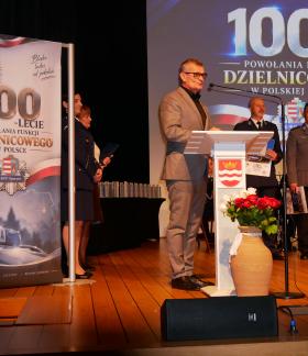 Burmistrz Drzewicy w szarym garniturze i okularach przemawia przy białej mównicy ozdobionej herbem oraz wazonem z czerwonymi różami. Po obu stronach stoją zaproszeni goście oraz nagrodzeni policjanci trzymający dyplomy i pamiątki. W tle widoczny jest baner oraz ekran z hasłem „100-lecie powołania funkcji dzielnicowego w Polskiej Policji”.