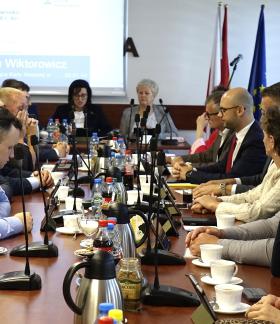 Sala konferencyjna UM w Drzewicy, w której odbywa się posiedzenie. Przy długim stole, na którym ustawiono mikrofony, filiżanki, butelki z napojami oraz dzbanki, siedzą po obu stronach radni i uczestnicy obrad. Na dalszym planie, na przedzie sali, zasiadają członkowie Prezydium Rady Miejskiej. Za nimi ekran projekcyjny oraz flagi.   
