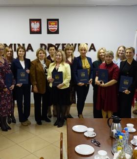 Pamiątkowe zdjęcie grupowe uczestników uroczystości. W sali konferencyjnej Urzędu Miejskiego w Drzewicy stoi grupa kilkunastu osób, elegancko ubranych w stroje wizytowe i garnitury. Większość osób trzyma w rękach ciemnoniebieskie pamiątkowe teczki z herbem Drzewicy. Wszyscy pozują przodem do aparatu.