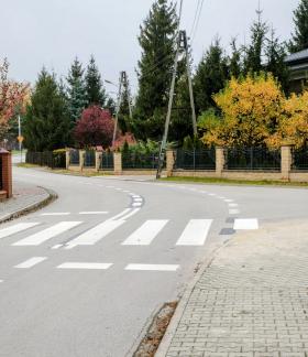 Przejście dla pieszych na łuku asfaltowej drogi z pylonami i znakiem D-6 w otoczeniu jesiennej zieleni i domów jednorodzinnych.