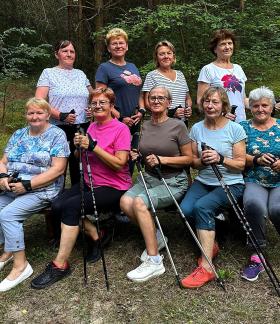 Grupowe zdjęcie dziewięciu uśmiechniętych kobiet w dojrzałym wieku, pozujących na tle leśnej zieleni. Pięć kobiet siedzi na drewnianej ławce, a cztery stoją tuż za nimi. Wszystkie panie ubrane są w wygodne, kolorowe stroje sportowe i trzymają kije do nordic walking.