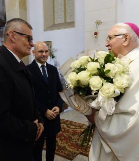 Arcybiskup Wacław Depo w białych szatach liturgicznych i różowej piusce przyjmuje duży bukiet białych róż wewnątrz kościoła. Naprzeciw niego stoi Burmistrz Drzewicy w ciemnym płaszczu i okularach, a obok niego Wiceburmistrz Drzewicy w garniturze.