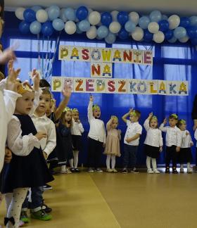 Grupa dzieci w odświętnych strojach z kolorowymi opaskami na głowach bierze udział w uroczystości pasowania na przedszkolaka. W tle napis „PASOWANIE NA PRZEDSZKOLAKA” oraz dekoracja z balonów. 