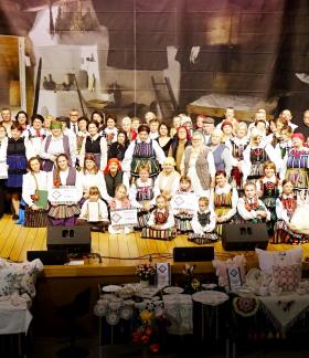 Zdjęcie grupowe uczestników Rewii na scenie sali widowiskowej RCK. W centrum osoby w tradycyjnych strojach ludowych, dzieci oraz goście. Na pierwszym planie stoły nakryte haftowanymi obrusami z wyrobami rękodzieła i poduszkami dekoracyjnymi. W tle dekoracja sceniczna z motywem dawnej chaty.