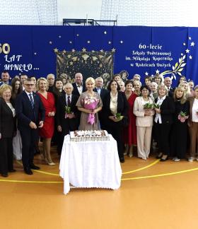 Pozowane zdjęcie grupowe kilkudziesięciorga uczestników jubileuszu 60-lecia SP w Radzicach Dużych. Przed grupą stoi stolik z uroczystym tortem, a w tle znajduje się niebieska ścianka dekoracyjna. 
