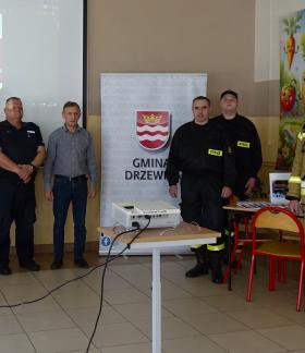 Grupa sześciu mężczyzn (policjanci, strażacy, cywil) stoi w rzędzie w sali prelekcyjnej między dwoma banerami reklamowymi a ekranem projekcyjnym.