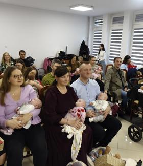 Grupowe zdjęcie uczestników spotkania w sali USC. Rodzice z niemowlętami na rękach oraz małe dzieci siedzą w krzesłach i prezentują otrzymane maskotki misiów oraz pamiątkowe teczki. 