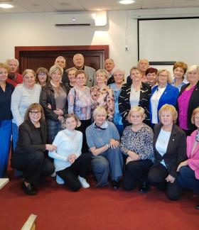 Pamiątkowe zdjęcie grupy uczestników i organizatorów spotkania rad seniorów, ustawionych w kilku rzędach w sali konferencyjnej. 