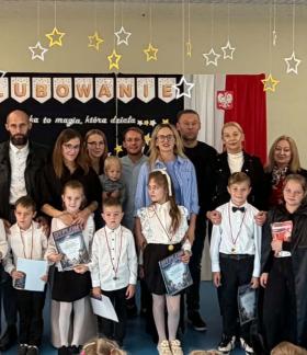 Szkolna uroczystość ślubowania. Zdjęcie grupowe pierwszoklasistów, ich rodziców, starszych uczniów oraz nauczycieli i gości na tle dekoracji z napisem „ŚLUBOWANIE”. 