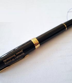  Eleganckie, czarne wieczne pióro ze złotą stalówką i złotymi wykończeniami, leżące na białym dokumencie papierowym. Na korpusie pióra znajduje się grawerowany napis w kolorze złotym: „Burmistrz Drzewicy Janusz Reszelewski”. W tle widoczny jest fragment podpisu i pieczątki na dokumencie.