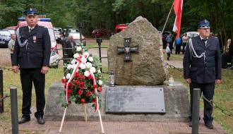 Warta honorowa dwóch druhów OSP przy obelisku w lesie „Parchowiec”. W tle las, samochody i kilkoro mieszkańców.