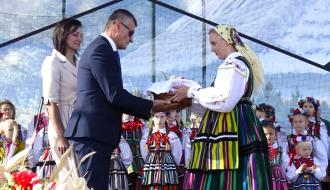 Na scenie Burmistrz Drzewicy Janusz Reszelewski odbiera bochen chleba od dożynkowej starościny Marty Jakubczyk. Obok Burmistrza stoi  Przewodnicząca Rady Miejskiej w Drzewicy Dorota Wiktorowicz