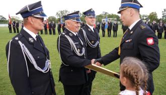 Zastępca Komendanta Wojewódzkiego PSP w Łodzi w obecności dziewczynki w stroju ludowym wręcza Prezesowi OSP Radzice akt włączenia jednostki do KSRG. Po bokach Prezesa Burmistrz Drzewicy i Naczelnik OSP Radzice. 