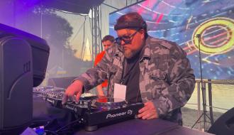 DJ w okularach przeciwsłonecznych i moro kurtce miksujący muzykę na scenie na tle telebimu. 