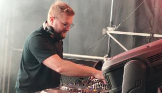 Mężczyzna w okularach i słuchawkach na szyi obsługujący konsolę dj-ską na scenie. 
