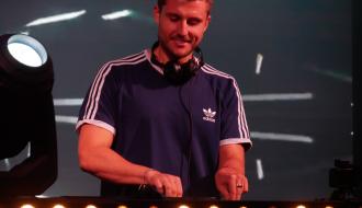 DJ w niebieskiej koszulce z białymi paskami stojący przy konsolecie dj-skiej na oświetlonej scenie. 