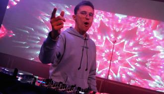 Młody mężczyzna w bluzie z kapturem stojący za konsolą dj-ską i wykonujący gest dłonią w stronę publiczności. 