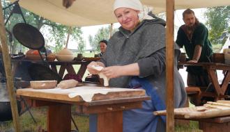 Kobieta w dawnym stroju wyrabiająca ciasto na drewnianej stolnicy pod zadaszeniem.