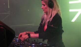 Kobieta w blond włosach i czarnej kurtce obsługująca konsolę dj-ską w zielonym świetle scenicznym. 