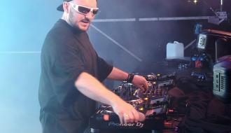 DJ w czarnej czapce z daszkiem i białych okularach przeciwsłonecznych grający na konsolecie na scenie.