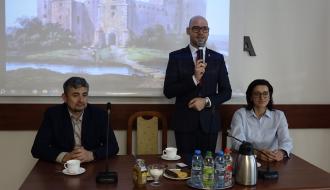 Przy stole prezydialnym na tle zdjęcia drzewickiego zamku w Drzewicy – od lewej: siedzi Zastępca Dyrektora RCPS w Łodzi Piotr Pietrzak, przemawia stojąc Zastępca Burmistrza Drzewicy Dominik Niemirski, siedzi Przewodnicząca Rady Miejskiej w Drzewicy Dorota Wiktorowicz.