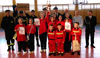 Młodzieżowe Drużyny Pożarnicze i Dziecięca Drużyna Pożarnicza z OSP Zakościele wraz z opiekunami prezentują zdobyte trofea, stojąc w hali sportowej.