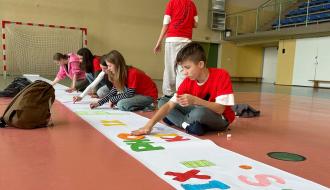 Sześcioro uczniów siedzi na parkiecie hali sportowej, wykonując baner Pho3nix Kids