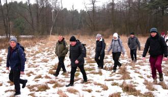 Siedmioosobowa grupa piechurów przemierza pokrytą śniegiem łąkę. W tle las