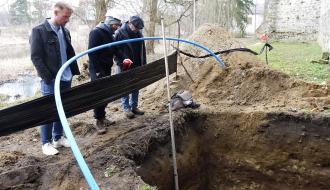 Trzech mężczyzn stoi przy wykopie archeologicznym i obserwuje jego przekrój, obok widoczna jest miarka głębokości oraz ściana zamku