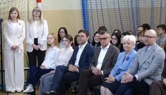 Grupa osób siedzi i stoi w sali gimnastycznej SP w Brzustowcu. W pierwszym rzędzie goście Forum siedzą na krzesłach. Obok nich znajdują się uczniowie, głównie młodzież, ubrani odświętnie. W tle widoczna jest siatka sportowa oraz fragment bramki do piłki ręcznej. Atmosfera jest oficjalna, a uczestnicy skupieni na przebiegu wydarzenia.