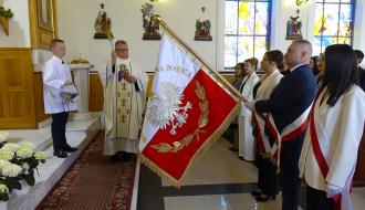 Wnętrze kościoła podczas uroczystości. Ksiądz święci sztandar w barwach biało-czerwonych z wizerunkiem orła. Obok ministrant trzymający kropidło i naczynie z wodą. Sztandar podtrzymuje poczet sztandarowy – kilka osób ubranych odświętnie.