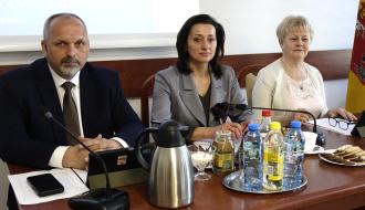 Trzy osoby z Prezydium Rady Miejskiej – mężczyzna i dwie kobiety - w formalnych strojach siedzą przy stole konferencyjnym z mikrofonami i dokumentami. Przed nimi znajdują się napoje i kawa. W tle widoczny fragment ekranu projekcyjnego oraz flaga.