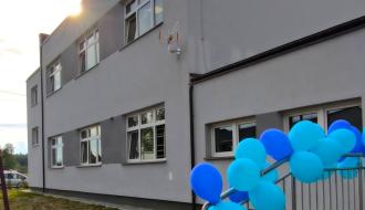 Nowo wybudowany, piętrowy budynek o szarej elewacji. Po prawej stronie widoczne są barierki schodów ozdobione niebieskimi i błękitnymi balonami. W tle świeci słońce.