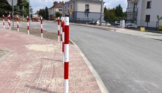 Nowo wybudowany chodnik z czerwonej kostki brukowej biegnący wzdłuż asfaltowej jezdni obok skrzyżowania dróg. Chodnik oddzielony od jezdni krawężnikiem oraz rzędem biało-czerwonych słupków połączonych łańcuchem. W tle budynki mieszkalne i zaparkowane samochody.