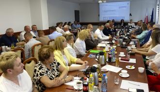 Uczestnicy sesji Rady Miejskiej w Drzewicy siedzący przy długim stole konferencyjnym. W tle widoczny ekran z programem obrad, na którym wyświetla się nazwisko Burmistrza Janusza Reszelewskiego. Obok flagi Polski i Unii Europejskiej.