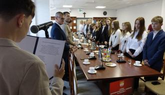 Młody radny czyta tekst ślubowania do mikrofonu podczas I Sesji Młodzieżowej Rady Miejskiej w Drzewicy. W tle, przy stole konferencyjnym, stoją pozostali radni oraz zaproszeni goście.