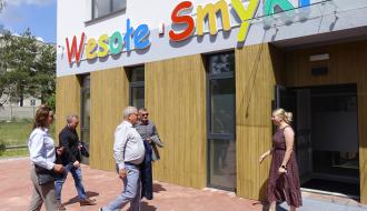 Grupa osób rozmawia przed wejściem do nowego budynku żłobka z kolorowym napisem „Wesołe Smyki”.