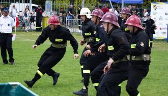 Dynamiczne ujęcie ze sportowo-pożarniczych zawodów na trawiastym boisku. Męska drużyna OSP Drzewica w czarnych mundurach i metalowych hełmach biegnie, mijając zbiornik z wodą widoczny w lewym dolnym rogu. W tle sędzia w białej koszuli, siatka ogrodzeniowa oraz zgromadzona publiczność.