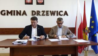Członek Zarządu Woj. Łódzkiego Artur Bagieński i Burmistrz Drzewicy Janusz Reszelewski siedzą przy drewnianym stole w urzędzie i podpisują oficjalne dokumenty. W tle, na białej ścianie, widnieje napis „GMINA DRZEWICA”, godło Polski oraz herb Drzewicy, a po prawej stronie stoją flagi państwowe i unijne.
