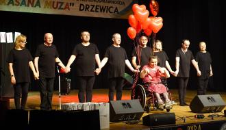 Na scenie grupa z WTZ w Drzewicy. Osiem osób w czarnych strojach stoi w rzędzie, trzymając się za ręce. Po prawej stronie na wózku inwalidzkim siedzi kobieta, trzymająca w dłoniach parę czerwonych butów. Nad nią unosi się pęk czerwonych balonów w kształcie serc. W tle transparent z nazwą przeglądu. 