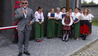 Mężczyzna w szarym garniturze i okularach przeciwsłonecznych przemawia do mikrofonu. Za nim stoi grupa kobiet w ludowych strojach oraz dziewczynka w tradycyjnym, pasiastym stroju opoczyńskim. Wszyscy stoją na kostce brukowej przed nowoczesnym budynkiem z ciemnej cegły. W tle widoczna jest biało-czerwona wstęga.