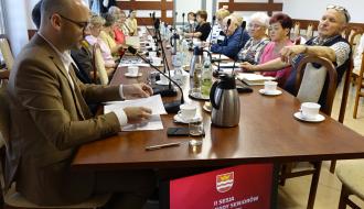Mężczyzna w jasnobrązowym garniturze siedzi na czele długiego stołu konferencyjnego i przegląda dokumenty podczas posiedzenia Miejskiej Rady Seniorów w Drzewicy. Przy stole siedzą starsze osoby, a pod blatem widoczny jest ekran z napisem „II SESJA MIEJSKIEJ RADY SENIORÓW W DRZEWICY, 17 czerwca 2026 r.”.