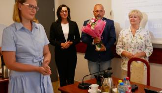 Cztery osoby stojące w sali konferencyjnej UM w Drzewicy podczas sesji rady Miejskiej. Na pierwszym planie po lewej Pani Dyrektor Przedszkola Samorządowego w Drzewicy w błękitnej sukience. W głębi Wiceprzewodniczący Rady trzyma bukiet różowych kwiatów, a obok niego stoją Przewodnicząca i Wiceprzewodnicząca Rady.