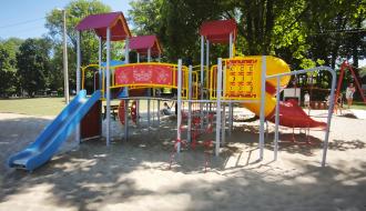 Kolorowy, rozbudowany zestaw sprawnościowy ze zjeżdżalniami i siatką wspinaczkową na piaszczystym placu zabaw w otoczeniu drzew